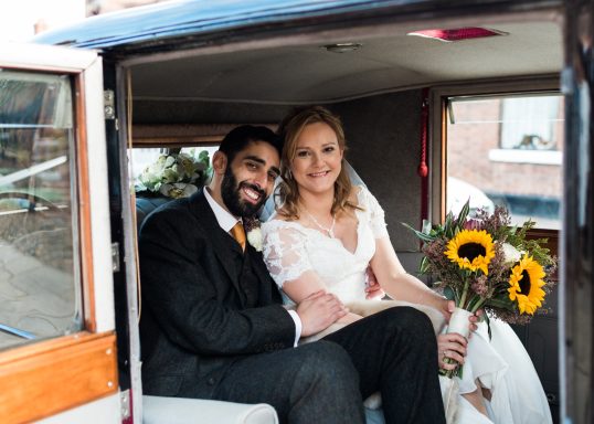 Beautiful couple sitting in wedding car with a bunch of sunflowers. The bride is radiant and happy, showing off her bridal makeup