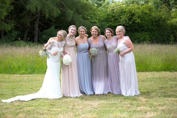 A bride with her six bridesmaids, standing in a line smiling at the camera. Bridal party makeup with glowing skin and natural bridal beauty.