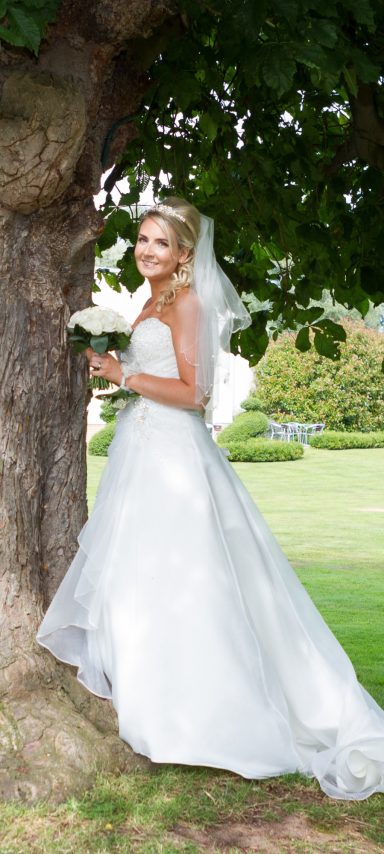 Smiling bride at Thornton Hall Hotel wedding after getting ready at home in Chester. Standing underneath a tree, holding a bouquet.